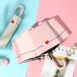 Parapluie rose ouvert avec motifs cœurs, plié à côté d'accessoires tendance, idéal pour rester chic sous la pluie.