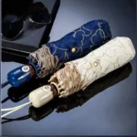 Parapluie automatique cœur élégant, disponible en bleu motifs dorés et beige, posé sur surface réfléchissante avec lunettes en fond.