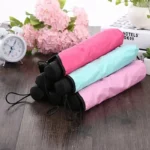 Parapluie femme coloré plié en rose et bleu sur table en bois, fleurs et horloge en arrière-plan, idéal pour un style élégant.