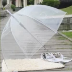 Parapluie transparent avec poignée blanche de la collection [Collection], posé sur un chemin en bois près d'un tapis et de magazines.