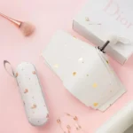 Parapluie cœur femme avec accessoires chic sur fond rose : pochette cœurs dorés, boîte luxe, boucles d'oreilles et pinceau tendance.