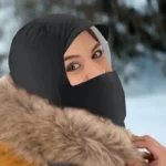 Femme souriante avec bonnet noir, cache-cou et manteau en fourrure, portant une capuche de pluie 3 en 1, dans un décor neigeux.