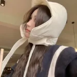 Femme coiffée d'un chapeau beige rembourré, vêtue d'une veste noire à rayures blanches, dans un intérieur cosy et chaleureux.