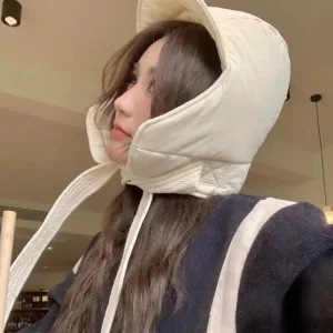 Femme coiffée d'un chapeau beige rembourré, vêtue d'une veste noire à rayures blanches, dans un intérieur cosy et chaleureux.