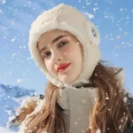 Femme souriante en chapka beige dans la neige, sous ciel bleu, portant une combinaison chaude avec capuche de pluie intérieur doux.