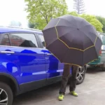Homme avec parapluie XXL noir coutures jaunes, près voiture bleue sous la pluie, entouré d'arbres, style [Collection], résistant.
