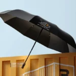 Parapluie noir pliable anti-retournement, design moderne avec logo 'No.Three', suspendu au-dessus d'un conteneur orange élégant.