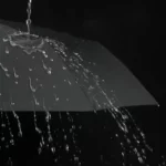 Parapluie homme pliant noir, résistant sous une pluie intense, gouttes d'eau visibles pour illustrer son efficacité et sa robustesse.