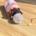 Parapluie luxe rose plié, motif floral élégant, embout strass étincelant, accessoire raffiné sur surface boisée.