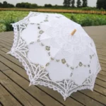 Parapluie mariage en dentelle blanc élégant, posé sur un sol en bois, avec un arrière-plan nature verdoyante et romantique.