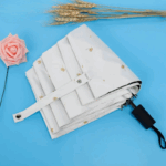 Parapluie plié blanc avec motifs dorés, sangle de maintien, rose rose et tiges de blé sur fond bleu.