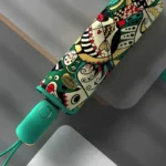 Parapluie pliant coloré avec motifs artistiques, design vibrant et fond turquoise, idéal pour un style unique et fonctionnel.