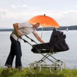 Parapluie poussette orange à clipser, homme l'ajustant au bord d'un lac pour offrir une protection solaire optimale à son enfant.