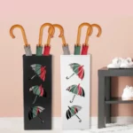 Porte parapluie en métal moderne noir et blanc avec parapluies colorés, fond rose pastel et chaussures blanches à côté.