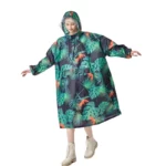 Femme avec poncho long motif tropical, capuche et poches, parfaite pour la pluie. Élégant et pratique, look tendance.