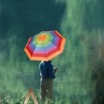 Chapeau parapluie arc-en-ciel porté par un pêcheur en plein air, entouré d'une nature verdoyante, idéal pour se protéger de la pluie.
