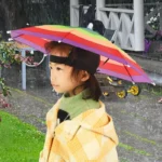 Enfant portant un chapeau parapluie coloré de la collection [Collection], idéal pour se protéger de la pluie avec style.