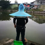 Homme avec chapeau parapluie pliant bleu au bord d'un plan d'eau, prêt à pêcher, avec bâtiments en arrière-plan.