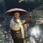 Homme souriant tenant une canne à pêche sous un chapeau parapluie uni, poisson dans filet, bord d'un lac ensoleillé
