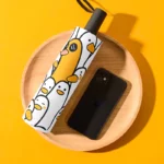 Parapluie rigolo avec motifs de canards mignons, sur plateau en bois à côté d'un téléphone noir, fond jaune lumineux.