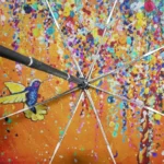 Parapluie automatique coloré avec motifs abstraits, gouttes vibrantes et colibri sur fond orange, design moderne et artistique.