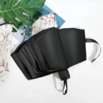 Parapluie femme pliable noir élégant, ouvert sur fond blanc avec feuilles tropicales, parfait pour allier style et praticité par temps de pluie.