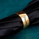 Parapluie homme luxe replié, anneau doré gravé Damun, design élégant sur fond bleu turquoise texturé.