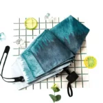 Parapluie plié bleu dégradé, style élégant, posé sur une surface à carreaux avec citrons et glaçons, idéal contre la pluie.