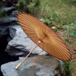 Parapluie japonais élégant en bambou avec toile marron, posé sur des rochers près d’un cours d’eau dans un cadre naturel verdoyant