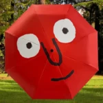 Parapluie rouge souriant, design unique avec yeux blancs et sourire noir, sur fond nature. Accessoire joyeux pour jours de pluie.