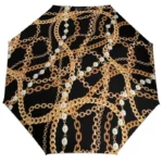 Parapluie pliant glamour noir avec motifs chaînes dorées et perles, alliant élégance et praticité pour vos journées pluvieuses.