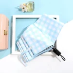 Parapluie pliable motif vichy bleu clair avec étui noir, posé sur fond pastel aux côtés d’un parfum et d’une trousse élégante.