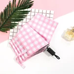 Parapluie vichy pliable rose et blanc sur fond blanc, accompagné d'une feuille verte et d'un flacon de parfum en arrière-plan.