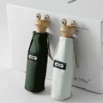 Parapluies grenouille stylisés avec poignées bois, yeux en plastique et housses vertes, exposés sur boîte avec texte imprimé.
