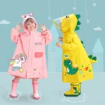 Deux enfants souriants en ponchos colorés, rose licorne et jaune dinosaure, prêts pour la pluie avec leurs bottes assorties.