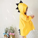 Poncho bébé dinosaure jaune avec capuche et détails noirs, idéal pour la pluie, enfant à côté de plantes décorées d'ananas.