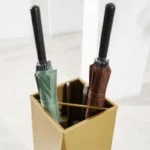 Porte-parapluie design doré, collection [Collection], tenant des parapluies verts et marrons avec des tiges noires apparentes.