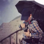 Homme avec parapluie noir et sac à dos, ajustant son équipement sur une balustrade en extérieur. Porte-parapluie sac à dos.