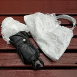 Parapluie mariage champêtre noir compact avec housse en dentelle blanche sur fond en bois, alliant élégance et utilité.