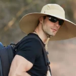 Jeune homme avec chapeau de pluie large bord anti-UV imperméable, lunettes de soleil, parfait pour activités extérieures ensoleillées.