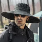 Homme avec chapeau pare-soleil anti-UV à large bord, lunettes de soleil et sac à dos, dans un cadre boisé, protection pluie et soleil.