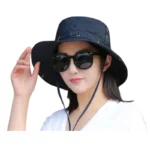 Femme souriante avec chapeau bob imperméable anti-UV noir, lunettes de soleil, main dans les cheveux, idéale contre la pluie.