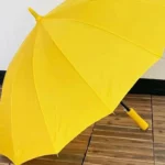 Parapluie droit jaune lumineux, ouvert sur un sol en bois, alliant style et durabilité pour affronter la pluie avec éclat.