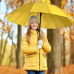 Fille souriante en manteau jaune avec un grand parapluie droit jaune, automne, sous les arbres aux feuilles dorées.