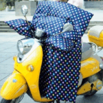 Scooter jaune avec tablier à motifs points blancs, protection élégante contre la pluie, stationné sur un trottoir urbain.