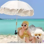 Parapluie poussette parasol beige à franges pour bébé, parfait pour la plage, avec un chien blanc et une mer turquoise en arrière-plan.