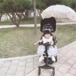 Petite fille dans une poussette avec un parapluie beige à franges, style parasol, dans un parc verdoyant et charmant.