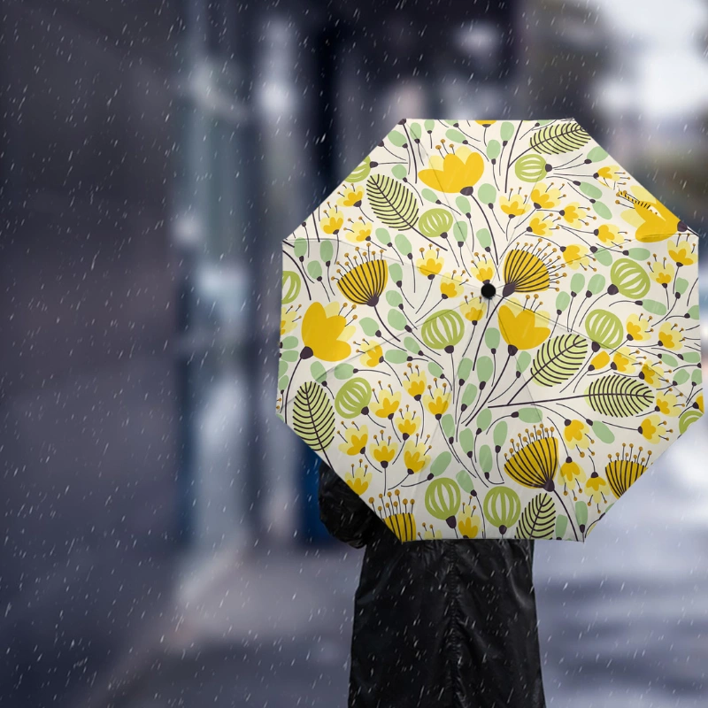 Parapluie automatique jaune et vert fleuri sous la pluie, tenu en main avec un arrière-plan urbain. Idéal pour toutes vos sorties.