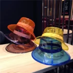 Chapeau de pluie transparent en PVC, coloris orange, jaune, bleu, tendance et pratique, exposé sur une table en bois.