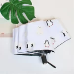 Parapluie beige automatique pliable avec motifs d'animaux polaires (pingouins, ours blancs) sur fond blanc, poignée noire élégante.